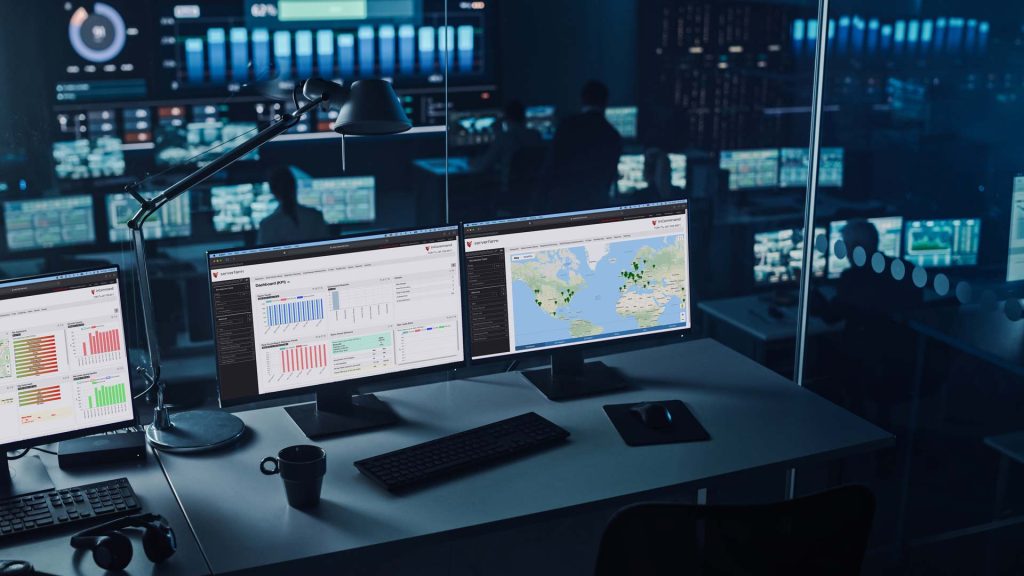 A high-tech control room with multiple monitors displaying data charts and maps, surrounded by dim lighting and people working in the background.