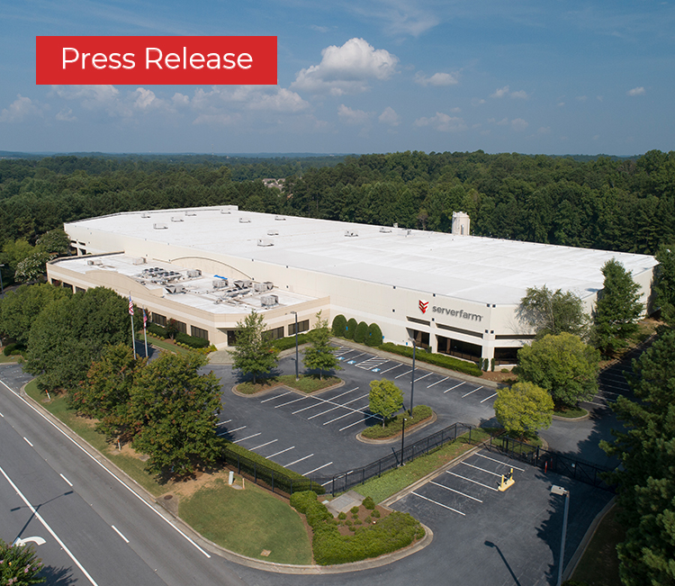 Aerial view of a large commercial building surrounded by trees and a nearly empty parking lot under a clear blue sky.
