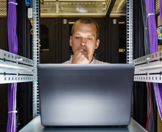 A person focuses intently on a laptop, surrounded by server rack equipment and colorful network cables.