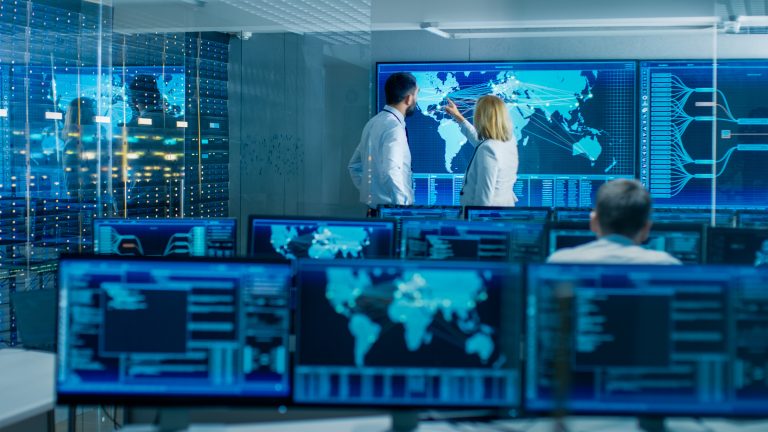 A modern control room with several screens displaying data, where two individuals discuss information on a large map.