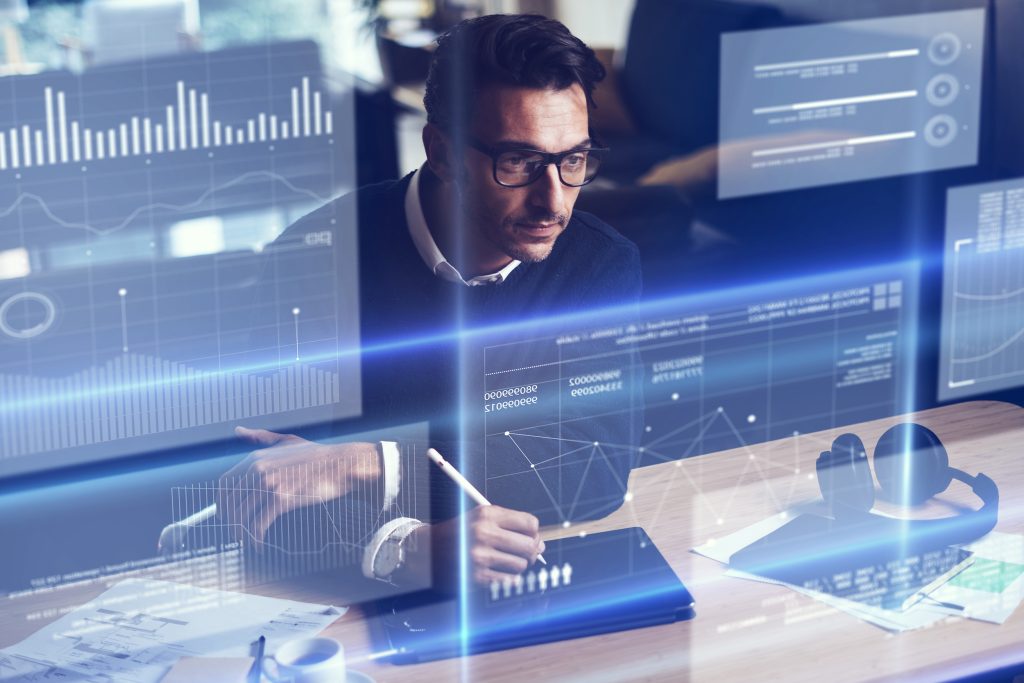 A man in glasses analyzes data on digital screens while taking notes in a modern office environment.