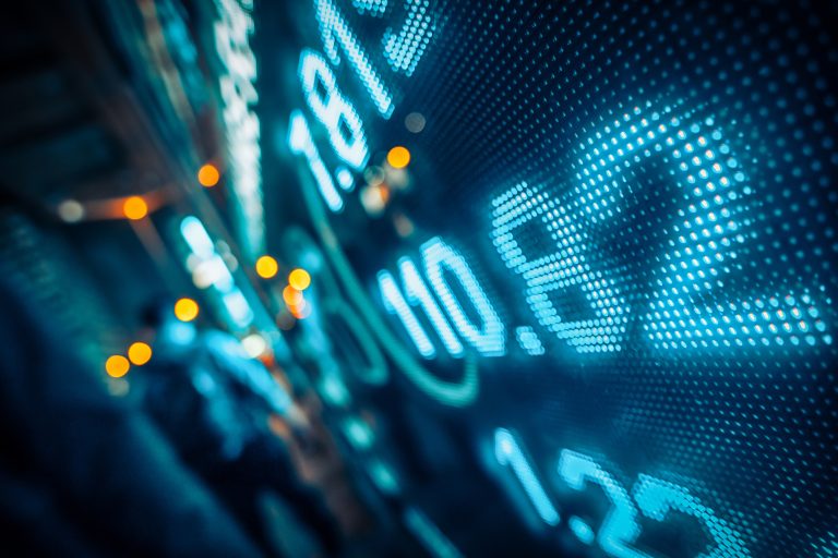 Illuminated digital financial numbers with a blurred background of figures and lights, suggesting a bustling trading environment.