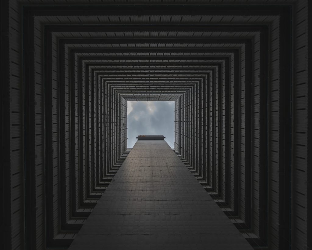 A geometric view looking up through a tall building's interior, showcasing a symmetrical pattern of repeating lines and shapes, with a glimpse of the sky above.