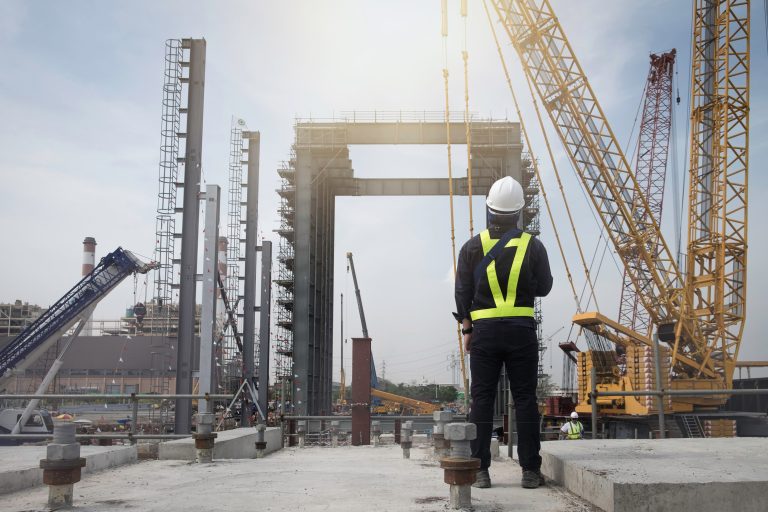 A construction worker in a reflective vest and helmet observes a large construction site with cranes and scaffolding under a bright sky.