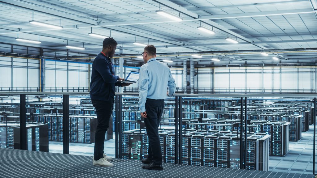 Two professionals discussing in a large data center filled with rows of server racks.
