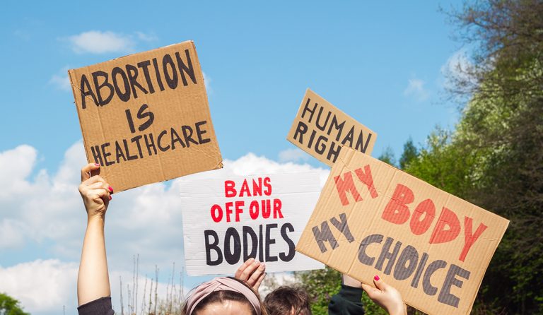 Protesters hold signs advocating for abortion rights, emphasizing themes of healthcare, human rights, and bodily autonomy against a blue sky backdrop.