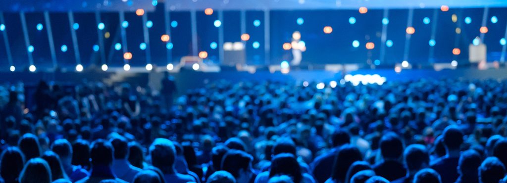 A large crowd at an event under dim blue lighting, with a speaker visible in the background.