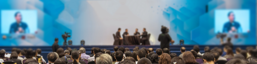 A large audience watches a panel discussion on stage, with speakers visible and screens displaying their images.