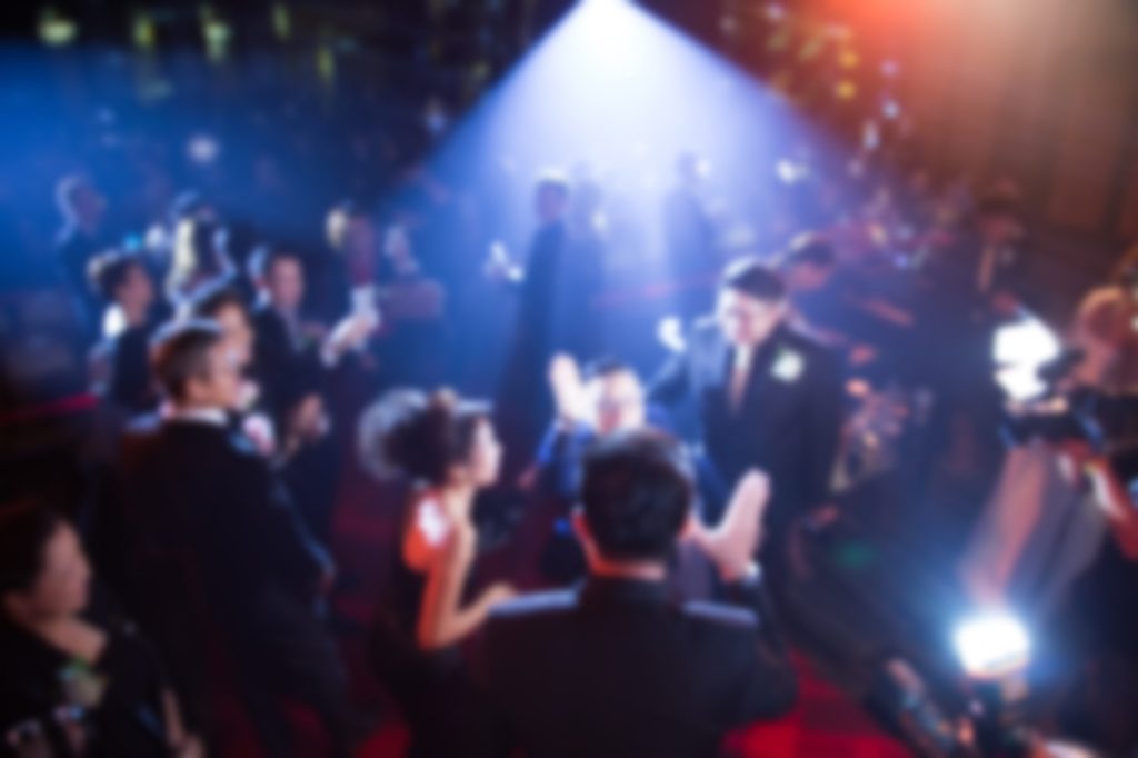 A celebration scene with attendees dressed in formal attire, illuminated by dramatic lighting and cameras capturing the moment.