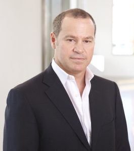 A man with short hair, wearing a dark suit and white shirt, poses confidently with a neutral expression in a modern setting.