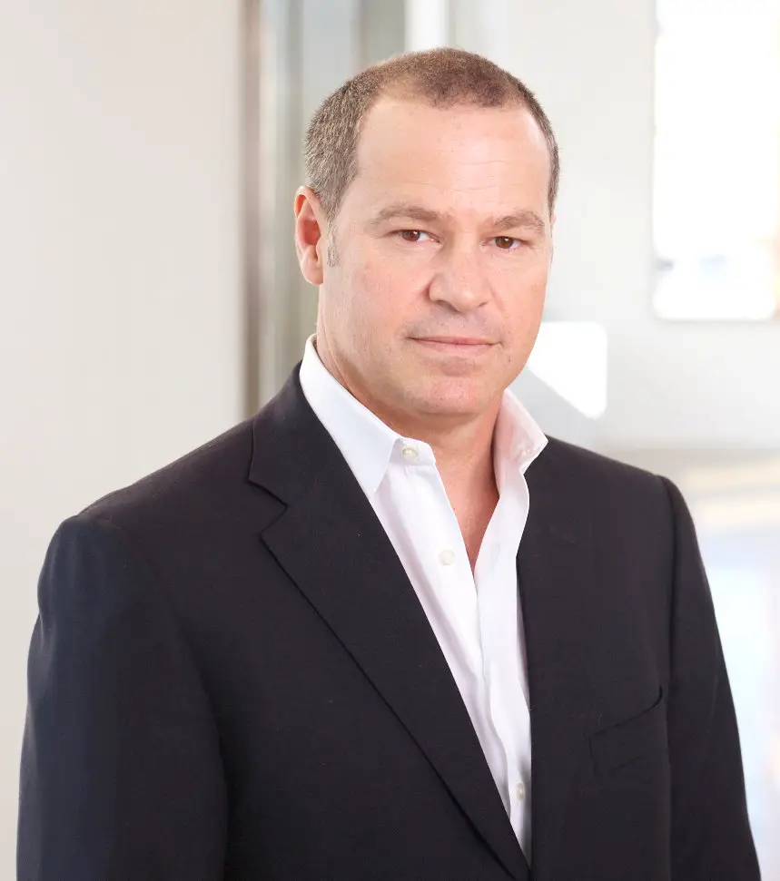 A man with short hair, wearing a dark suit and white shirt, poses confidently with a neutral expression in a modern setting.