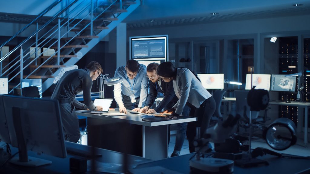 A group of professionals collaborate over a table in a dimly lit office, surrounded by multiple computer monitors displaying data.
