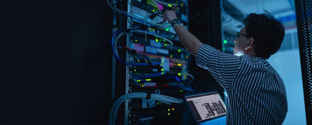 A technician works on server racks filled with equipment and illuminated indicators, with a laptop nearby in a dimly lit environment.