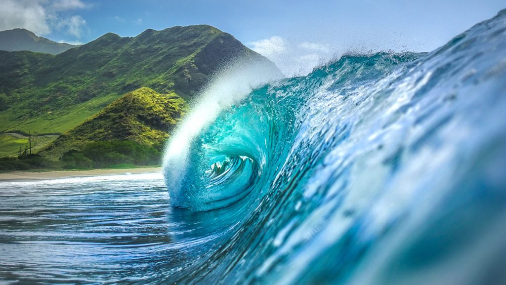 A stunning ocean wave curls with vibrant blue hues, set against a backdrop of lush green mountains and a clear sky.