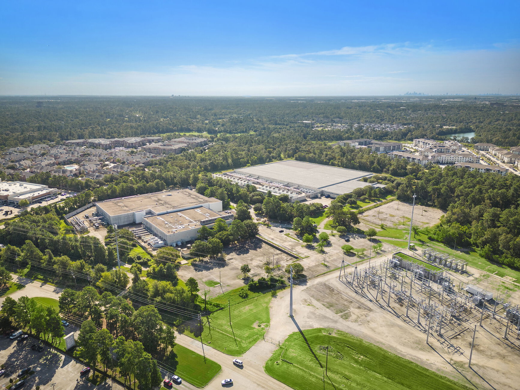 Aerial view of a suburban area featuring warehouses, green spaces, power lines, and residential neighborhoods under a clear blue sky.