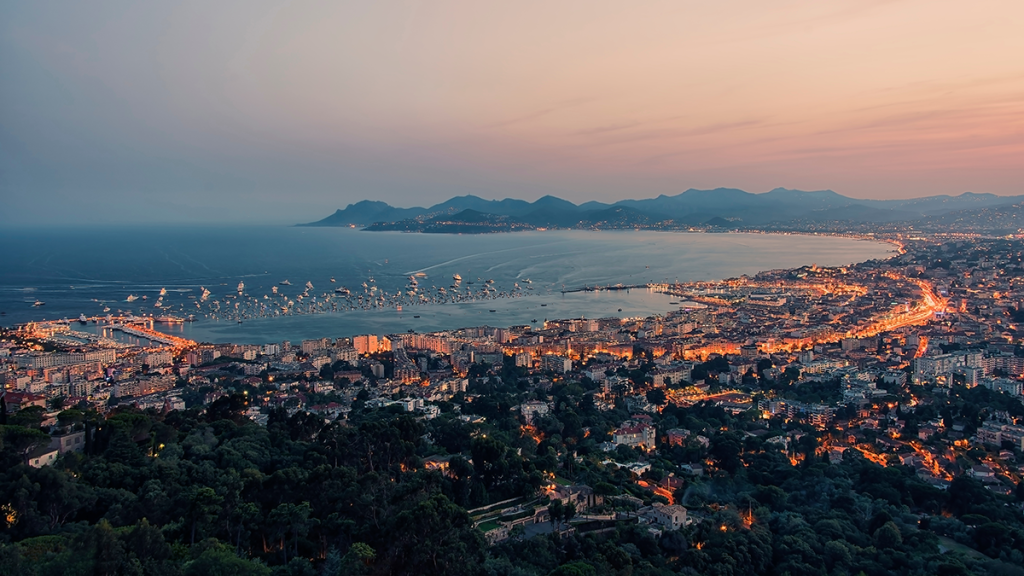 A panoramic view of a coastal city at sunset, featuring a lively harbor, hills in the background, and warm city lights.