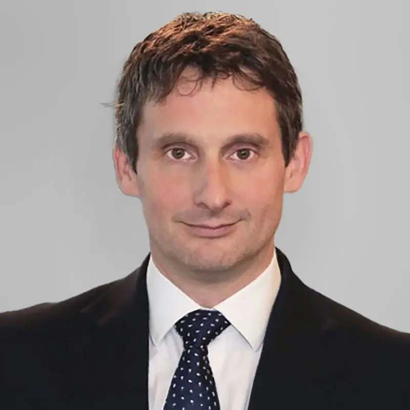A man in a black suit with a white shirt and polka dot tie, posing against a plain grey background.