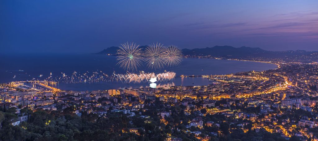 A vibrant evening view of a coastal city illuminated by fireworks over the ocean, showcasing a lively atmosphere and city lights.