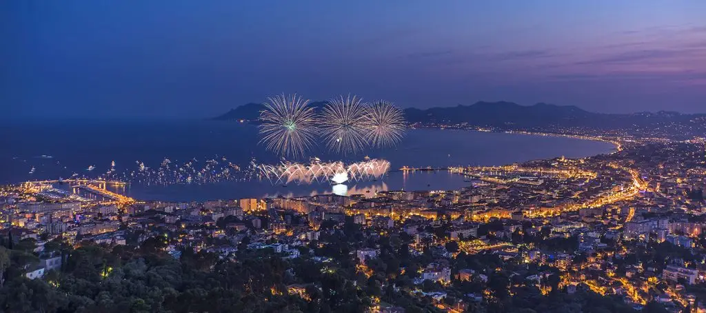 A vibrant evening view of a coastal city illuminated by fireworks over the ocean, showcasing a lively atmosphere and city lights.
