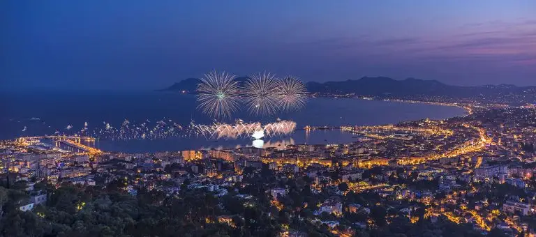 A vibrant evening view of a coastal city illuminated by fireworks over the ocean, showcasing a lively atmosphere and city lights.