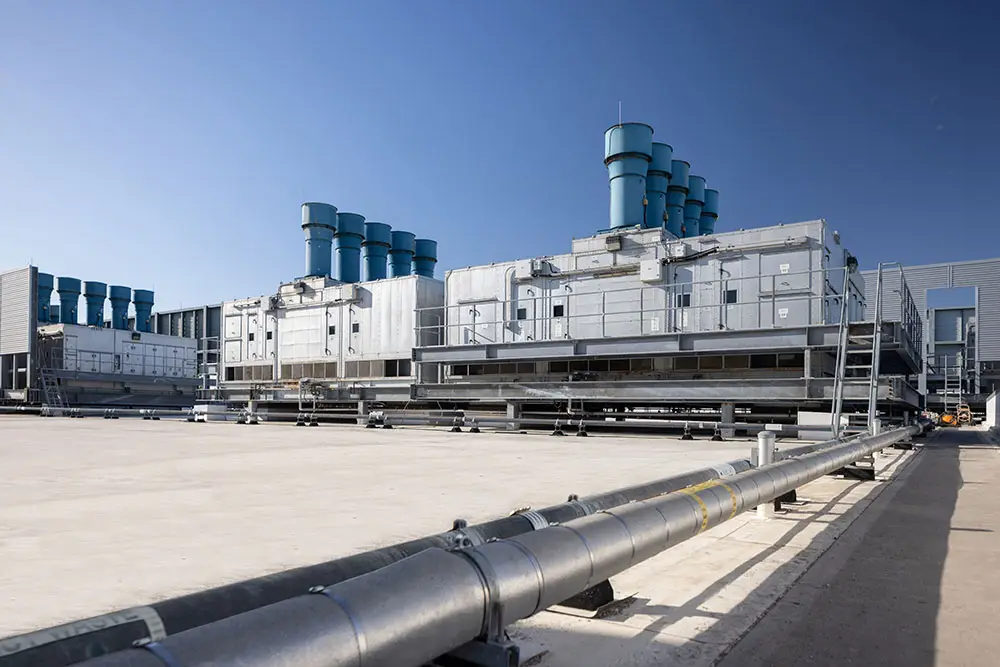 Industrial structures with large blue exhaust stacks, set against a clear blue sky, alongside a metal pipeline extending across the foreground.