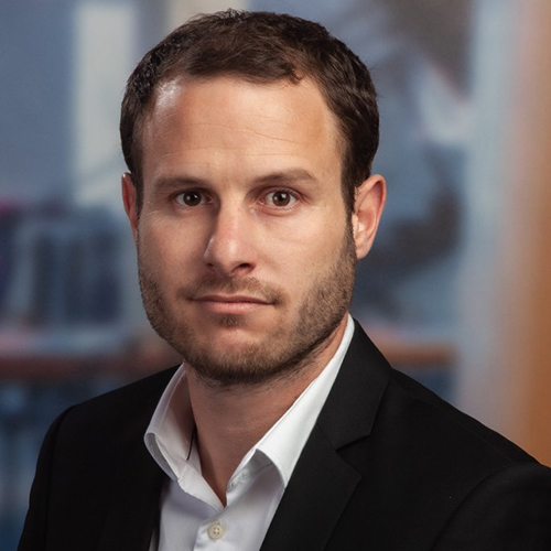 A man in a suit looks directly at the camera, displaying a serious expression in an office-like setting.