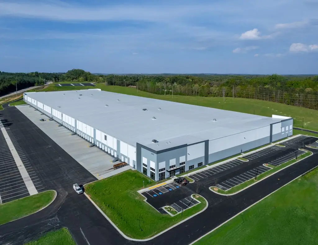 A large industrial building with a flat roof and ample parking space, set against a backdrop of greenery and clear blue sky.