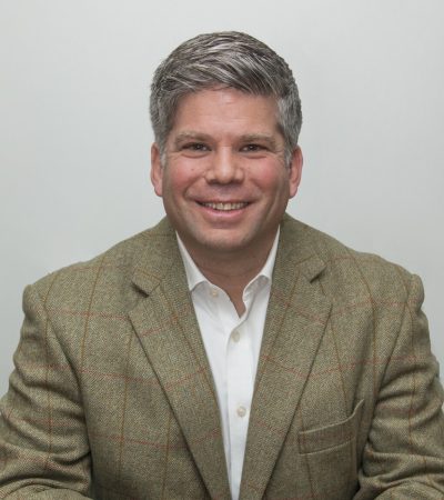 A casually dressed man in a tan blazer smiles at the camera, sitting against a light gray background.