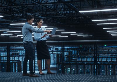 A man and a woman discuss data insights while using a laptop in a large, modern data center.