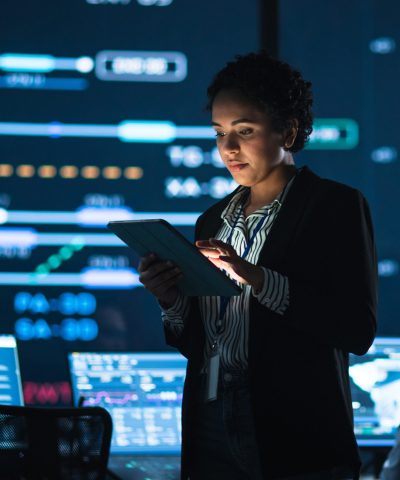 A woman in a dark environment interacts with a digital tablet, illuminated by glowing screens filled with data and analytics.