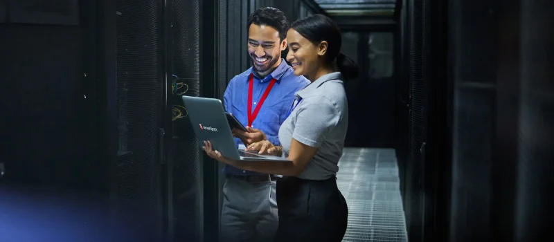 Two professionals collaborate in a server room, engaged with a laptop and discussing work while surrounded by server racks.