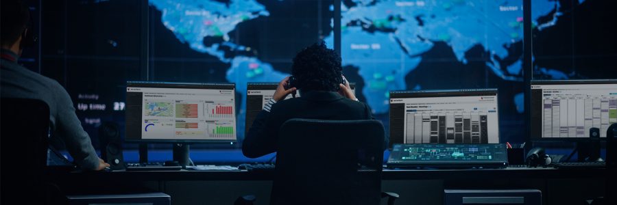 Two people are seated at computers in a control room, analyzing data on screens with a world map backdrop.
