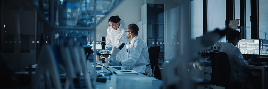 Two scientists in laboratory coats collaborate over a microscope, surrounded by modern lab equipment and a dimly lit, high-tech environment.