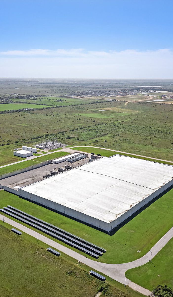 An aerial view of a large warehouse surrounded by expansive green fields, with clear blue skies in the background.
